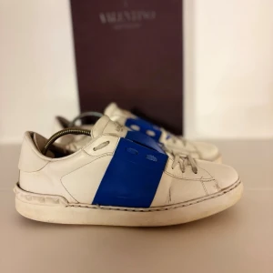 Valentino Open sneakers vit/blå - Säljer ett par Valentino Open sneakers i vitt skinn med bred blå rem över mitten. Klassisk låg modell med rund tå, snörning och subtila nitar på hälen. Perfekta för dig som vill ha en clean men ändå unik look. Medföljs box+ dustbag Skicket är som på bild 
