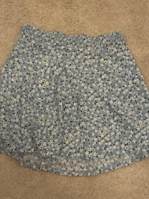 Blommig blå kortkjol med resår - Söt kortkjol i ljusblått med små vita och mörkblå blommor. Kjolen har en bekväm resår i midjan och är gjord i ett lätt och luftigt material, perfekt för varmare dagar. Passar dig som gillar en avslappnad och somrig stil.