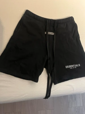Svarta shorts från Fear of God Essentials - Svarta shorts från Fear of God Essentials med logga tryckt på vänster ben. Modellen har snörning i midjan och sidofickor. Perfekta för en avslappnad och trendig stil. Tillverkade i mjuk bomull för maximal komfort.