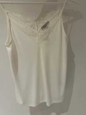 Vit spetsdetaljerad topp från  H&M storlek XS - Säljer en vit topp från H&M med tunna axelband och vacker spetsdetalj vid ringningen. Toppen har en feminin v-ringad form och är perfekt för lager på lager eller till jeans. Enkel och stilren design som passar till många outfits.