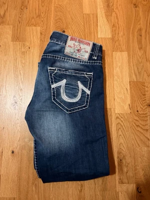 True Religion blå jeans med kontrastsömmar - Säljer ett par blå jeans från True Religion med tydliga vita kontrastsömmar och den klassiska hästsko-loggan på bakfickorna. Jeansen har normal passform och raka ben, tillverkade i slitstarkt denimtyg. Perfekta för dig som gillar streetwear och vill sticka ut. Priset är inte hugget i sten. 