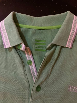 Grön pikétröja från BOSS Hugo Boss - Snygg ljusgrön pikétröja från BOSS Hugo Boss med klassisk krage och knäppning framtill. Tröjan har vita och rosa ränder på krage och ärmslut samt korta ärmar. Perfekt för en avslappnad men stilren look.