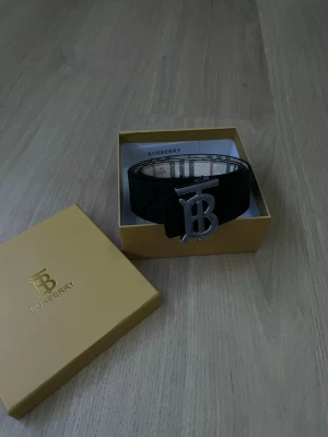 Svart Burberry bälte med silverdetalj - Snyggt svart bälte från Burberry med klassiskt rutigt innerfoder och stor silvrig BB-logga som spänne. Kommer i original gul Burberry-box. Perfekt accessoar för att lyfta din outfit med en lyxig touch.