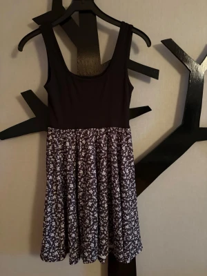 Svart blommig kortklänning - Söt kortklänning med svart överdel och blommigt mönstrad kjol i vitt och svart. Ärmlös modell med rundad halsringning och lätt utställd kjol. Aldrig använd!
