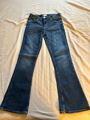 Blå bootcut jeans från Gina Tricot - Säljer ett par klassiska blå bootcut jeans från Gina Tricot i storlek 158. Jeansen har en snygg utsvängd passform, fem fickor och kontrastsömmar. Perfekta för dig som gillar retrovibbar och vill ha ett par bekväma jeans i bomull.