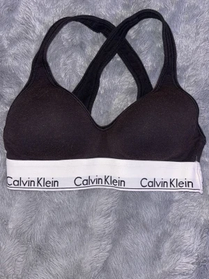 Svart sport-bh från Calvin Klein - Svart sport-bh från Calvin Klein med vadderade kupor och bred, vit resår med logga runt om. Bh:n har korsade axelband i ryggen och är tillverkad i mjukt, stretchigt material för extra komfort.