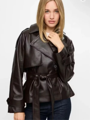 Brun skinnjacka med bälte - Snygg mörkbrun skinnjacka med bred krage och knytskärp i midjan. Jackan har stora slag, knappar framtill och långa ärmar med dekorativa detaljer vid ärmslut. Perfekt för dig som vill ha en trendig och stilren look.