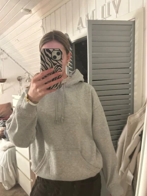 Grå basic hoodie med huva - En klassisk grå hoodie med huva och dragsko, samt en stor magficka framtill. Hoodien har ribbade muddar vid ärmslut och nederkant. Perfekt för en avslappnad och stilren look. Defekter syns på bild.