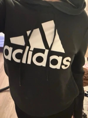 Svart Adidas hoodie med stor logga - Svart hoodie från Adidas med stor vit logga och klassiska ränder på bröstet. Tröjan har huva med dragsko och en avslappnad passform, perfekt för en sportig och casual stil.
