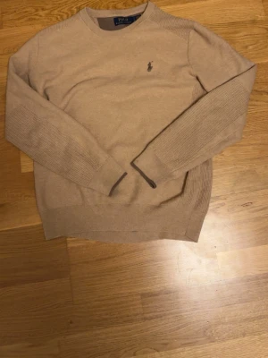Ralph lauren  - Det är en stickad tröja i en ljusbrun nyans med en rund halsringning. På vänster bröst sitter den välkända Ralph Lauren-loggan med ryttaren. Ärmarna har en distinkt textur med ribbstickade partier som löper längs med dem, och även nederkanten och ärmsluten är ribbstickade. Kragen har en mörkare brun kant på insidan.