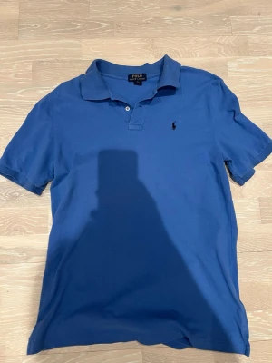 Blå pikétröja från Polo Ralph Lauren - Pikétröja från Polo Ralph Lauren med färgen harbor island blue. Pikén har krage, två knappar vid halsen och korta ärmar. Tillverkad i mjuk bomull som känns skön mot huden. Köpt på zalando för 1695 mitt pris 400. Hör av dig vid minsta fundering 