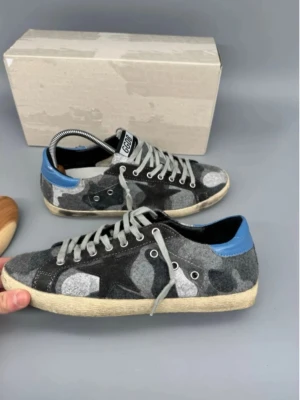 Grå camo sneakers från Golden Goose - Snygga sneakers från Golden Goose med grått och svart camouflage-mönster, blå häl och vita snören. Modellen heter Superstar och har en klassisk låg siluett med rund tå och platt sula. Ovandelen är i filtliknande material och detaljer i skinn.