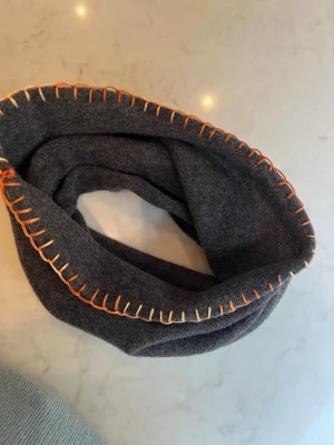 Buff / tubhalsduk med detaljer - Mysig mörkgrå tubhalsduk i merinoull med handsydd, dekorativ kantsöm. Perfekt för att hålla värmen och ge en cool detalj till din outfit. Enkel att dra över huvudet och sitter skönt runt halsen. Även toppen i skidbacken! 