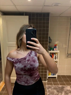 Mörklila one shoulder topp - Säljer en snygg mörklila one shoulder topp med batikmönster i lila och vitt. Toppen är tight och har en asymmetrisk design som ger en trendig look. Perfekt för dig som vill sticka ut lite extra. Använd en gång! Nypris 230kr