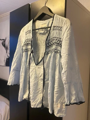 Ljusblå boho-blus från Odd Molly - Supersöt ljusblå blus från Odd Molly med broderade detaljer och smock upptill. Blusen har knytband vid halsen och svarta broderier längs ringningen och ärmsluten. Tillverkad i ett tunt, luftigt material som känns riktigt skönt.
