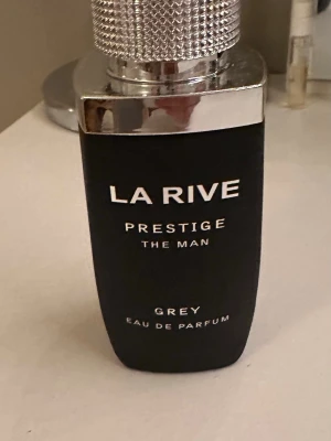 La Rive Prestige The Man Grey EdP - La Rive Prestige The Man Grey Eau de Parfum har en stilren svart flaska med silverdetaljer och tydlig logga. Parfymen är framtagen för män och flaskan har en modern, nästan ny har använt den två ggr bara 