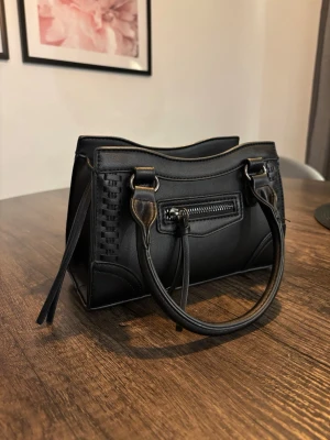 Svart handväska  - Svart handväska från Gina tricot. Väskan har ett stort fack med dragkedja upptill, med två små fack på insidan. Samt ett litet fack med dragkedja på framsidan av väskan. Det medföljer ett avtagbart axelband med justerbar längd. Väskan säljs inte längre på Gina tricot☺️ (Pris kan alltid diskuteras)