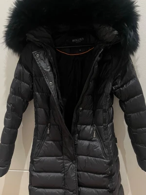 Svart lång dunjacka från Hollies - Snygg svart dunjacka från Hollies med lång passform och stor fluffig huva. Jackan har quiltad design, dragkedjor framtill och två fickor med dragkedja. Perfekt för kalla dagar och ger en riktigt stilren look.