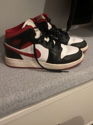 Nike Air Jordan 1 svart/röd/vit - Säljer ett par Nike Air Jordan 1 sneakers i svart, rött och vitt. Klassisk modell med snörning, rund tå och platt sula. Tillverkade i skinn med ikoniska detaljer och färgblock. Perfekta för dig som gillar streetwear och vill ha en tidlös sneaker.