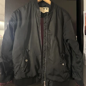 Svart bomberjacka från Junkyard - Svart bomberjacka från Junkyard med klassisk passform, ribbade muddar och dragkedja framtill. Jackan har snedställda fickor med tryckknapp och en ärmficka med dragkedja. Fodret är vinrött för en snygg kontrast. Perfekt för dig som vill ha en stilren och urban look.