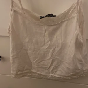 Vit spetskantad linnetopp Hollister - Säljer en söt vit linnetopp från Hollister med tunna axelband och fina spetsdetaljer längs både halsringning och nederkant. Toppen är croppad och har en lätt och luftig känsla, perfekt för varma dagar eller lager på lager.