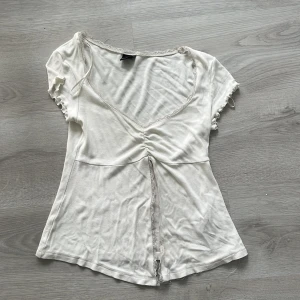 Vit v-ringad topp från Gina Tricot - Supersöt vit topp från Gina Tricot med v-ringning, spetsdetaljer längs kanterna och rynk framtill. Korta ärmar med volangkant och en feminin passform. Perfekt att styla med jeans eller kjol för en trendig look. Knappt använt😊