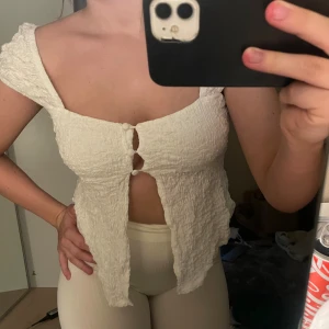 Vit croppad blus med knappar - Supersöt vit croppad blus med smockat tyg och två klädda knappar framtill. Toppen har korta ärmar, fyrkantig ringning och öppen front. Perfekt för dig som gillar romantisk och trendig stil.