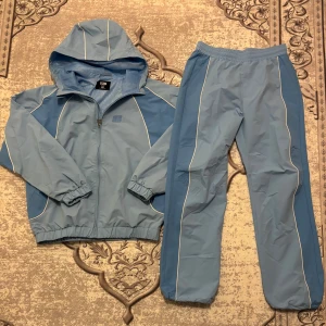 Blå vindjacka från FSBN med huva - Säljer en blå vindjacka från FSBN med huva och vita detaljer längs sömmarna. Jackan har dragkedja framtill, elastiska muddar och meshfoder på insidan. Perfekt för dig som gillar sportig stil och vill ha en lätt jacka med snygga linjer. Har tyvärr inte prislappen kvar, har bara använt den 3-4 gånger. Köpte för 999 kr. Vid frågor skicka!  Byxorna är baggy så de blir S båda blir som S.