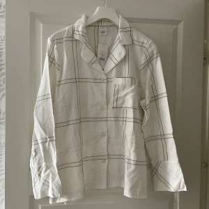 Vit rutig pyjamas i linne - Säljer en vit pyjamas i mjukt linne med rutigt mönster i svart. Skjortan har klassisk krage, bröstficka och knäppning framtill. Byxorna har resår i midjan och raka ben. Perfekt för dig som gillar en chill och stilren look.