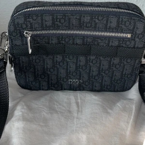 Svart axelväska från Dior - Snygg svart axelväska från Dior med klassiskt monogrammönster och bred justerbar axelrem. Väskan har silverfärgade dragkedjor och detaljer, samt en praktisk ytterficka framtill. Tillverkad i textil och skinn för en lyxig känsla. Box ingår men inget kvitto.                                                  