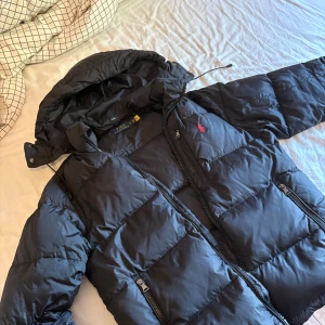 Svart pufferjacka från Polo Ralph Lauren - Svart pufferjacka från Polo Ralph Lauren med huva och dragkedjor framtill. Jackan har en röd broderad logga på bröstet, flera fickor med dragkedja och justerbar dragsko i huvan. Perfekt för kalla dagar och riktigt snygg streetstil. Pris kan även diskuteras😁passar som M/S