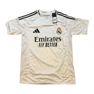Real Madrid vit matchtröja Adidas - Säljer en officiell Real Madrid fotbollströja från Adidas i vit färg med svarta detaljer och klubbmärke broderat på bröstet. Tröjan har korta ärmar, klassiska svarta axelränder och är tillverkad i lätt, ventilerande polyester med AEROREADY-teknologi.