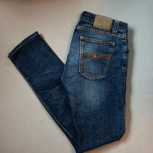 Blå jeans från Nudie Jeans, 30/32 - Snygga blå jeans från Nudie Jeans med klassisk orange söm på bakfickan och läderpatch i midjan. Modellen har slim ben och slimmad passform. Tillverkade i slitstarkt denim med cool tvättad look. Perfekta för dig som gillar stilrena och tidlösa jeans.