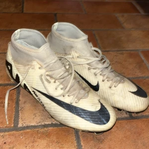 Nike Mercurial vita fotbollsskor - Säljer ett par Nike Mercurial fotbollsskor i vitt med svarta detaljer och snörning. Skorna har högre strumpliknande skaft och platt sula med dobbar för gräsplan. Tillverkade i syntetmaterial och designade för snabbhet på planen.