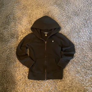 Svart hoodie från Polo Ralph Lauren - Svart hoodie från Polo Ralph Lauren med dragkedja framtill och klassisk vit logga broderad på bröstet. Tröjan har huva, känguruficka och är tillverkad i mjuk bomull. Perfekt för en chill och stilren look.