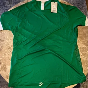 Hammarby fotbollströja från Craft - Säljer en officiell Hammarby fotbollströja från Craft i grönt och vitt med breda vertikala ränder. Tröjan har korta ärmar, vit krage och Hammarbys klubbmärke på bröstet. Materialet är lätt och andas, perfekt för match eller träning. Sponsorer tryckta på framsidan.