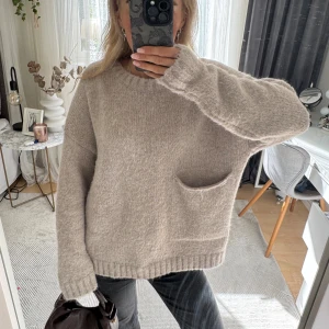 Beige stickad oversized tröja - Mysig beige stickad tröja med oversized passform och en stor ficka framtill. Tröjan har rund halsringning, långa ärmar och ribbade muddar. Perfekt för lager-på-lager och chill dagar.