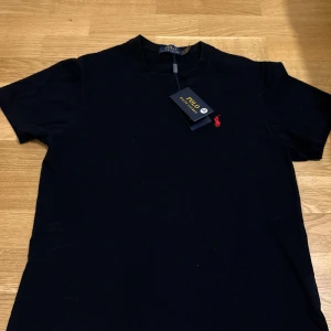 Svart t-shirt från Polo Ralph Lauren - Svart t-shirt från Polo Ralph Lauren med klassisk röd broderad logga på bröstet. T-shirten har rund hals och korta ärmar, tillverkad i mjuk bomull som känns skön mot huden. Perfekt för dig som gillar stilrena och ikoniska plagg.