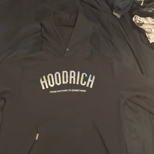 Svart hoodie från Hoodrich i storlek L - Svart hoodie från Hoodrich med stor silvrig logga och texten 'FROM NOTHING TO SOMETHING' på bröstet. Tröjan har huva med knappdetalj och en stor magficka framtill. Perfekt för dig som gillar streetwear och vill ha en clean look.