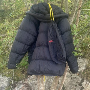 Svart dunjacka från Fjällräven, L - Säljer en svart, riktigt puffig dunjacka från Fjällräven med gula snören och huva. Jackan har två stora fickor med knappar, broderad logga på bröstet och är tillverkad i polyamid med dunfyllning. Perfekt för kalla dagar och har en loose passform.
