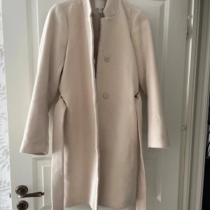 Beige kappa med bälte och knappar - Stilren beige kappa med rak passform, knappar framtill och ett matchande bälte i midjan. Jackan har lång ärm och enkel design utan krage, vilket ger en clean look. Perfekt för dig som gillar minimalistisk stil och vill ha något snyggt till höst och vinter.