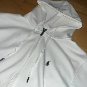 Vit hoodie från Polo Ralph Lauren - Säljer en stilren vit hoodie från Polo Ralph Lauren med dragkedja och huva. Tröjan har snörning i huvan, två fickor framtill och den klassiska broderade loggan på bröstet. Perfekt för en clean och avslappnad look.