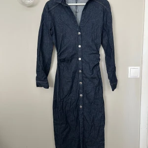 Mörkblå jeansklänning med knappar ZARA - Snygg lång jeansklänning i mörkblå denim med långa ärmar och knappar hela vägen framtill. Klänningen har klassisk skjortkrage och markerad midja. Perfekt för dig som gillar en stilren och tidlös look.