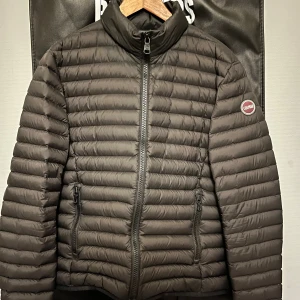 Svart dunjacka från Colmar, stl 48 - Snygg svart dunjacka från Colmar Originals i storlek 48. Jackan har horisontella sömmar, hög krage och två dragkedjeförsedda fickor framtill. Perfekt för kyliga dagar och har en clean, modern look med diskret logga på ärmen.