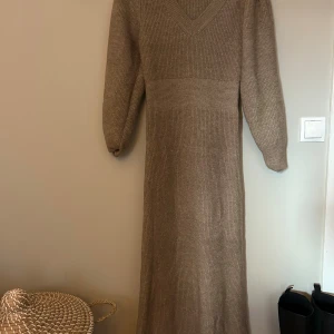 Beige stickad långklänning AWARE - Mysig beige stickad långklänning från AWARE med v-ringning och långa ärmar. Klänningen har en ribbad struktur och är perfekt för kyligare dagar. Enkel och stilren design som är lätt att matcha med boots eller sneakers.