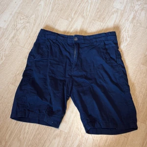 Mörkblå bomullsshorts med fickor - Snygga mörkblå shorts i bomull med klassisk passform. De har två sidofickor fram och en bakficka med knapp. Stängs med knapp och dragkedja framtill. Perfekta för varma dagar och har en enkel, stilren look.