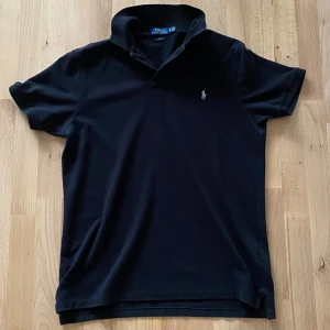 Svart Polo Ralph Lauren t-shirt XL - Svart t-shirt från Polo Ralph Lauren i slim fit-modell. Klassisk pikékrage och korta ärmar, med den ikoniska broderade loggan på bröstet. Tillverkad i mjuk bomull som känns skön mot huden. Perfekt för dig som gillar stilrena och tidlösa plagg.