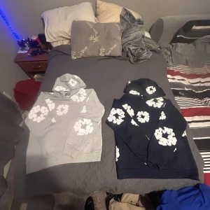 Denim Tears hoodie med blommor - Två hoodies från Denim Tears i storlek L, en grå och en marinblå, båda med vita blommiga tryck över hela tröjan. Klassisk hoodie-modell med huva och känguruficka, perfekt för dig som gillar streetwear och vill sticka ut med ett unikt mönster.