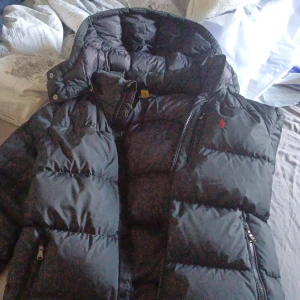 Svart dunjacka från Polo Ralph Lauren - Säljer en svart dunjacka från Polo Ralph Lauren med huva och dragkedja. Jackan har en klassisk pufferstil med två fickor framtill och diskret röd logga på bröstet. Perfekt för kalla dagar och riktigt snygg till streetwear.