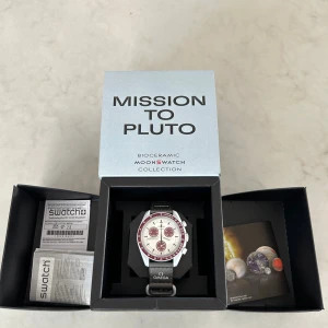 Omega x Swatch Mission to Pluto - Omega x Swatch Speedmaster Mission to Pluto från MoonSwatch-kollektionen. Klockan har en vit urtavla med vinröda detaljer och en vinröd bezel. Boetten är i biokeramik och armbandet är svart med Speedmaster- och Omega-logga. Baksidan har en bild på Pluto. Har endast använts ett fåtal gånger! Ser helt ny ut! Och inga skador! Pris kan diskuteras!